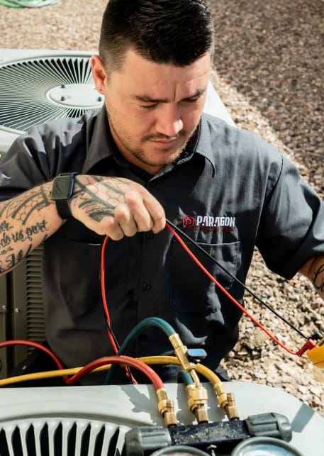 A tattooed technician from Paragon Service Pros repairs an air conditioner, showcasing skill and expertise in HVAC work.