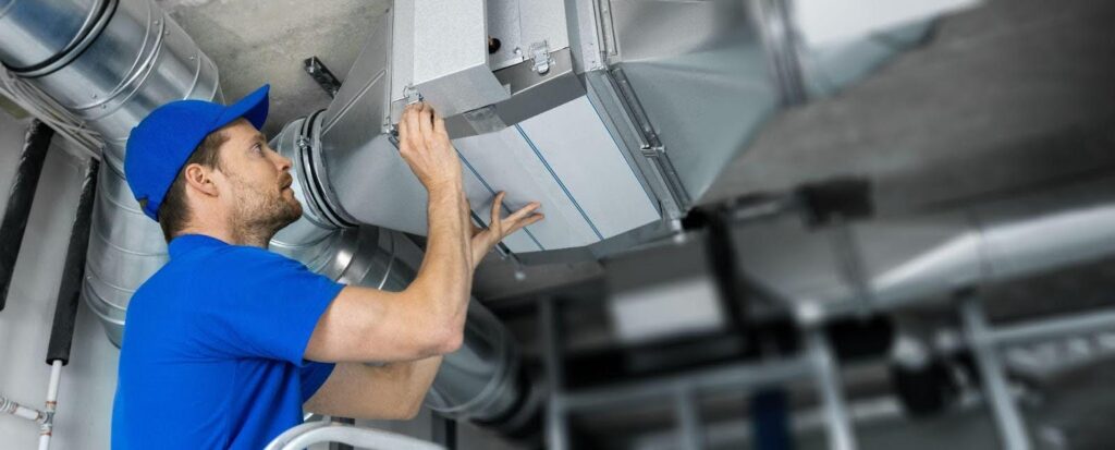 A man wearing a blue shirt and hat is repairing a ducted air conditioner, focused on his task